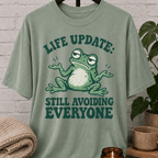 Funny introvert frog T-shirt featuring a shrugging frog and the phrase “Life Update Still Avoiding Everyone,” a sarcastic avoid people graphic tee for socially selective and low social battery humor fans.