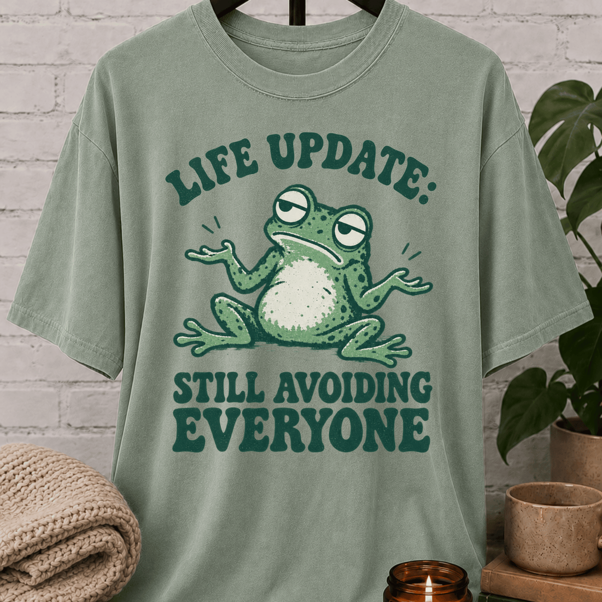 Funny introvert frog T-shirt featuring a shrugging frog and the phrase “Life Update Still Avoiding Everyone,” a sarcastic avoid people graphic tee for socially selective and low social battery humor fans.
