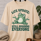 Funny introvert frog T-shirt featuring a shrugging frog and the phrase “Life Update Still Avoiding Everyone,” a sarcastic avoid people graphic tee for socially selective and low social battery humor fans.
