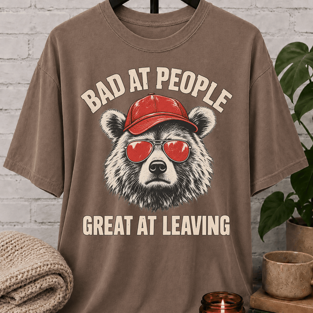 Socially awkward bear T-shirt featuring a bear in sunglasses and a red cap with the phrase “Bad at People Great at Leaving,” a sarcastic graphic tee for people who hate small talk and leave early.