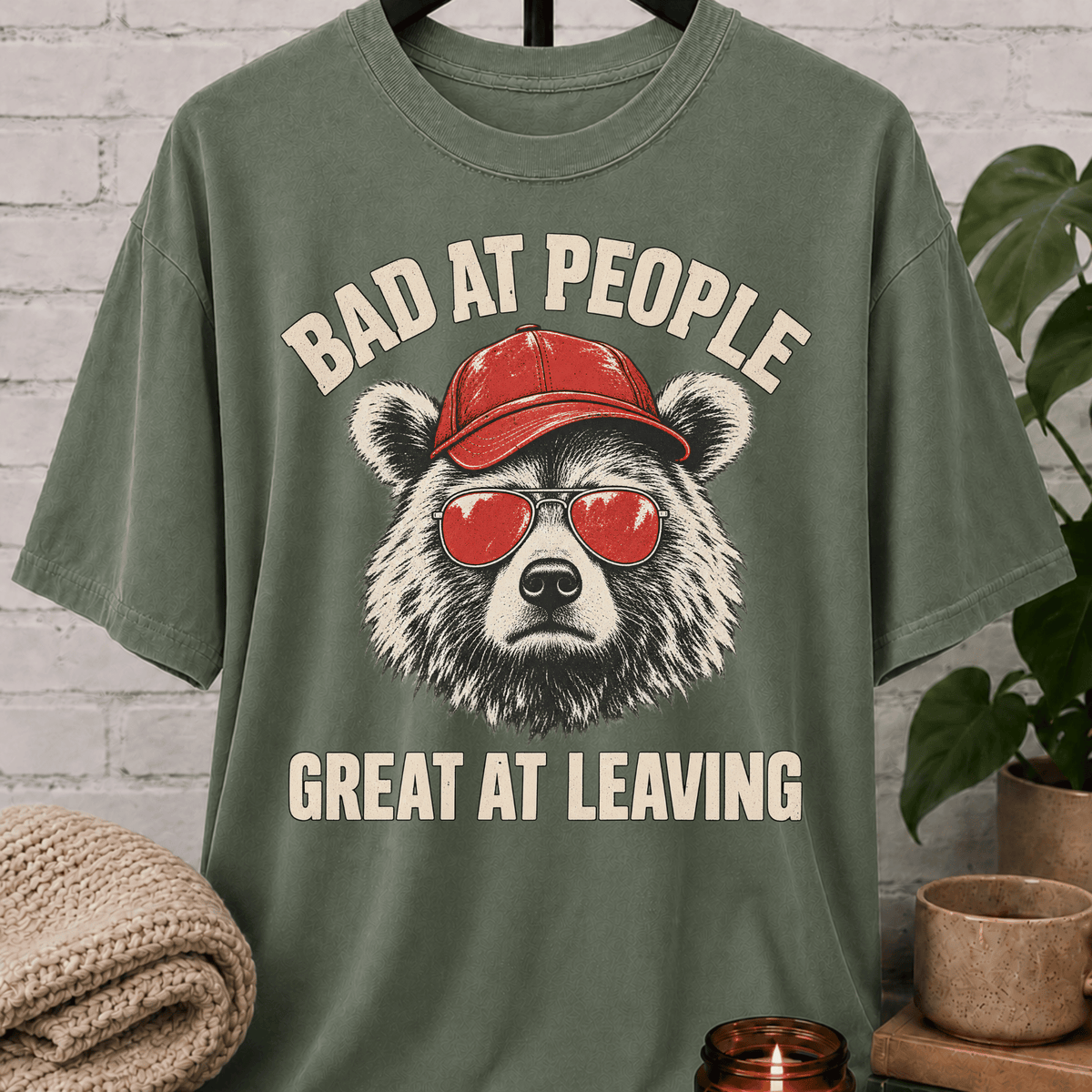 Socially awkward bear T-shirt featuring a bear in sunglasses and a red cap with the phrase “Bad at People Great at Leaving,” a sarcastic graphic tee for people who hate small talk and leave early.