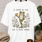 Coffee lover frog graphic T-shirt featuring a relaxed green frog holding a coffee mug among wildflowers with the phrase “I Could Go Out But I Have Coffee,” an indoorsy stay-home shirt for frog lovers and coffee fans.