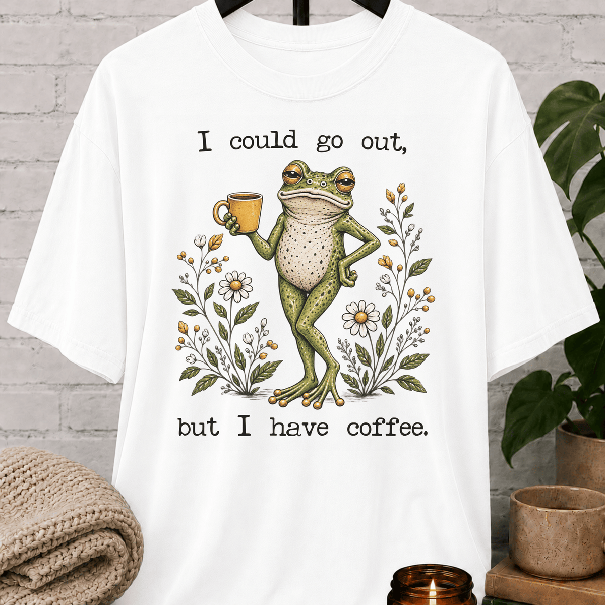 Coffee lover frog graphic T-shirt featuring a relaxed green frog holding a coffee mug among wildflowers with the phrase “I Could Go Out But I Have Coffee,” an indoorsy stay-home shirt for frog lovers and coffee fans.