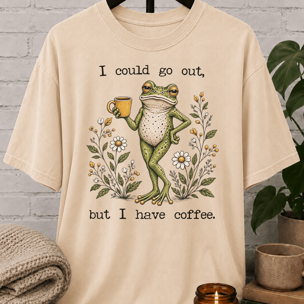 Coffee lover frog graphic T-shirt featuring a relaxed green frog holding a coffee mug among wildflowers with the phrase “I Could Go Out But I Have Coffee,” an indoorsy stay-home shirt for frog lovers and coffee fans.