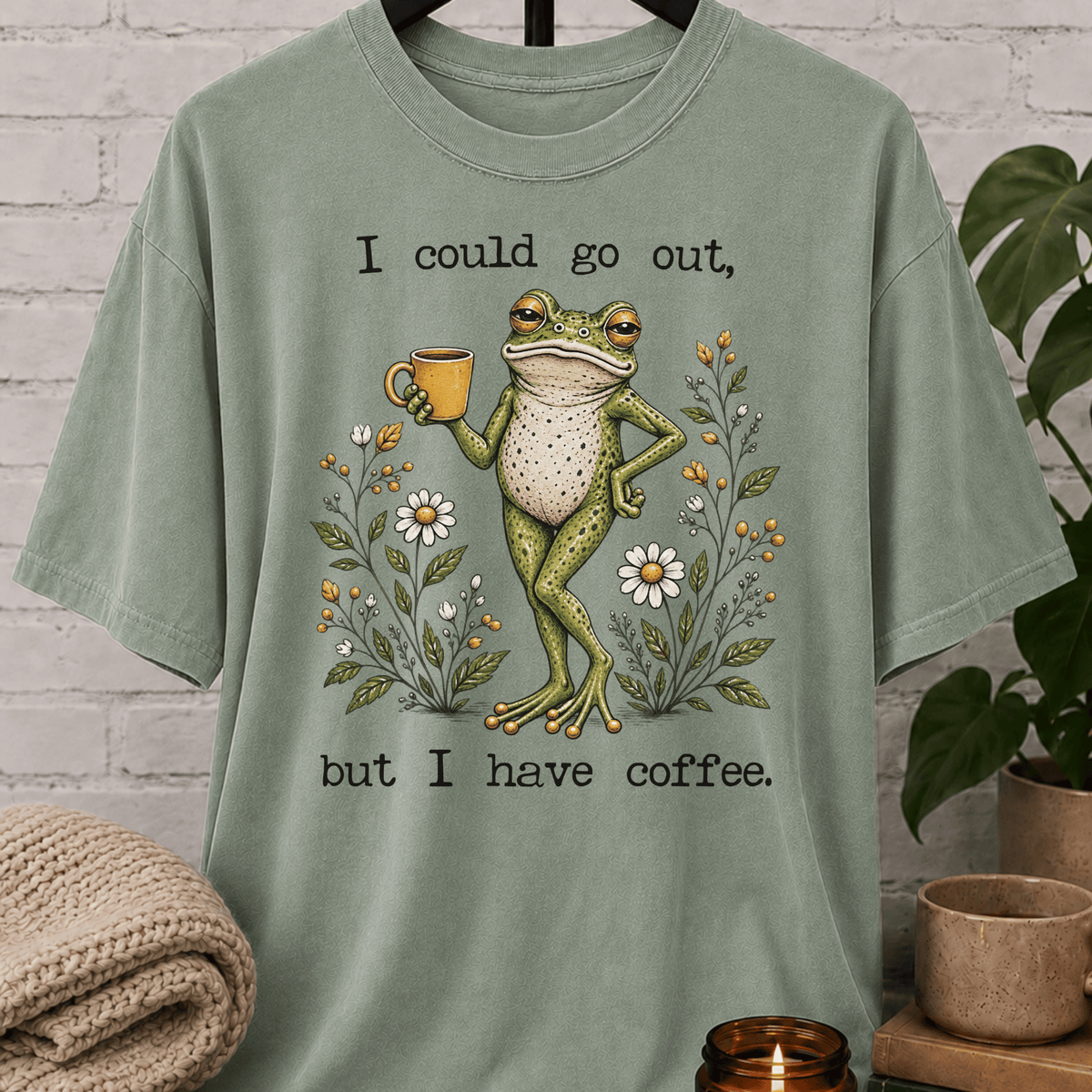 Coffee lover frog graphic T-shirt featuring a relaxed green frog holding a coffee mug among wildflowers with the phrase “I Could Go Out But I Have Coffee,” an indoorsy stay-home shirt for frog lovers and coffee fans.