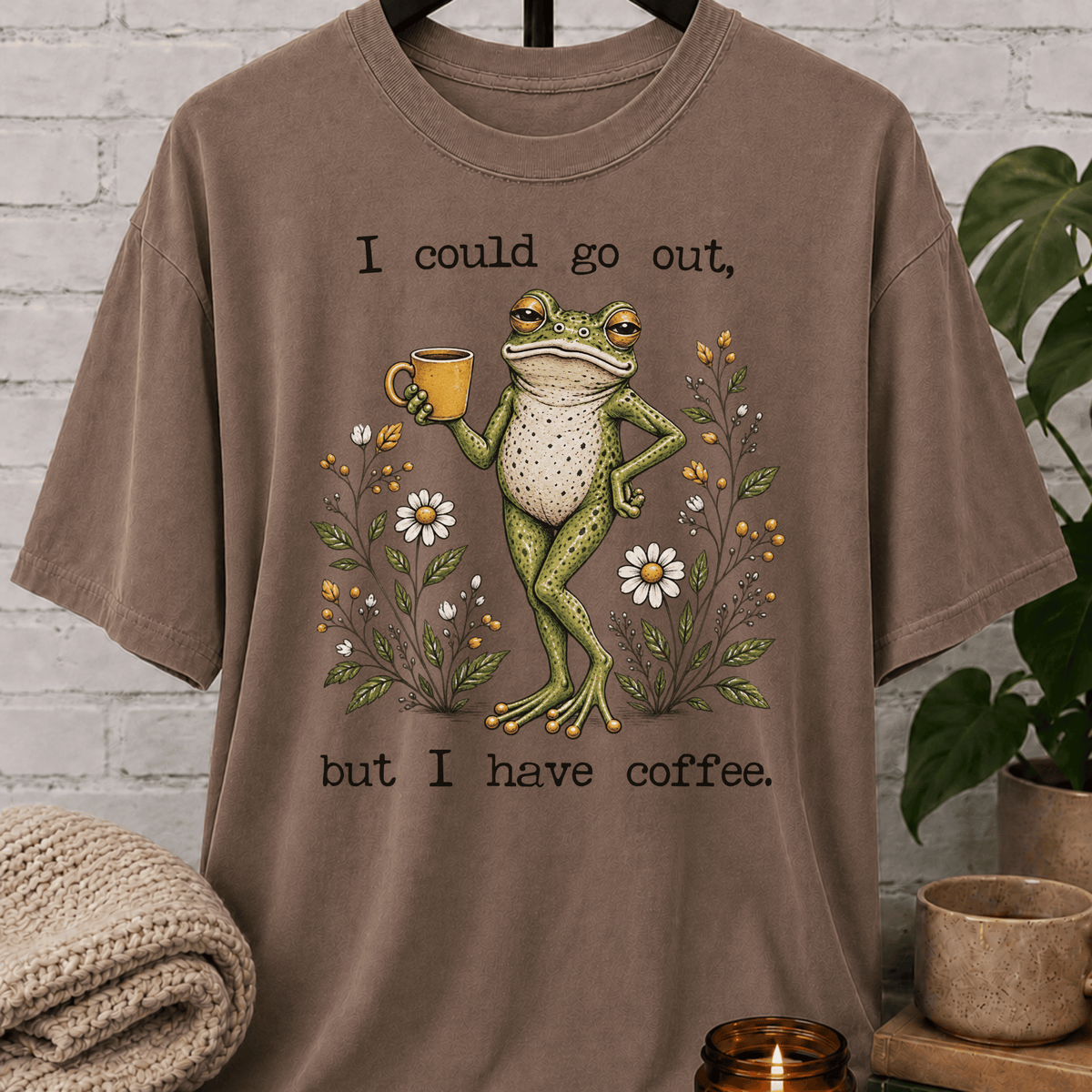 Coffee lover frog graphic T-shirt featuring a relaxed green frog holding a coffee mug among wildflowers with the phrase “I Could Go Out But I Have Coffee,” an indoorsy stay-home shirt for frog lovers and coffee fans.