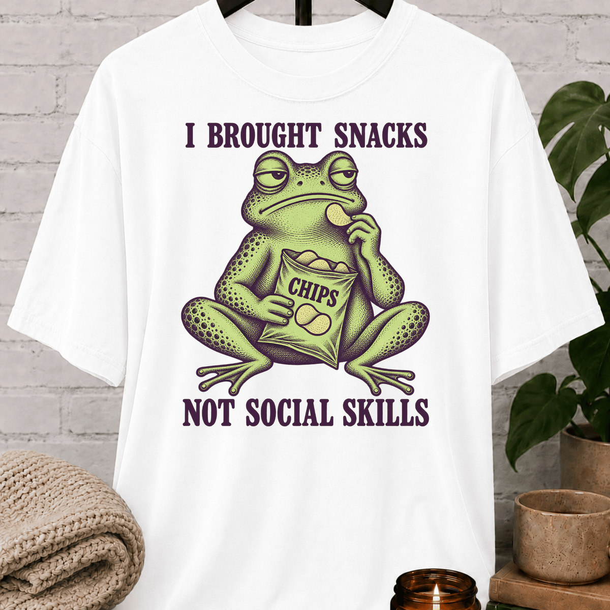 Socially awkward frog graphic T-shirt featuring an unimpressed green frog eating chips with the phrase “I Brought Snacks Not Social Skills,” a sarcastic snack humor shirt for awkward people and low social battery types.