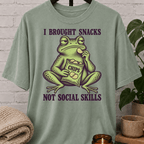 Socially awkward frog graphic T-shirt featuring an unimpressed green frog eating chips with the phrase “I Brought Snacks Not Social Skills,” a sarcastic snack humor shirt for awkward people and low social battery types.