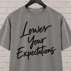 Lower Your Expectations graphic T-shirt featuring bold handwritten lettering, a blunt humor shirt for low-expectation personalities, sarcastic humor fans, and people who prefer keeping the bar low.