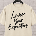Lower Your Expectations graphic T-shirt featuring bold handwritten lettering, a blunt humor shirt for low-expectation personalities, sarcastic humor fans, and people who prefer keeping the bar low.