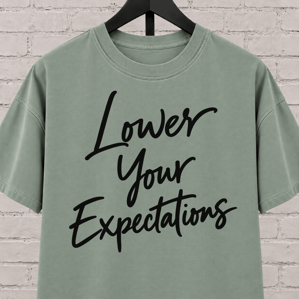 Lower Your Expectations graphic T-shirt featuring bold handwritten lettering, a blunt humor shirt for low-expectation personalities, sarcastic humor fans, and people who prefer keeping the bar low.