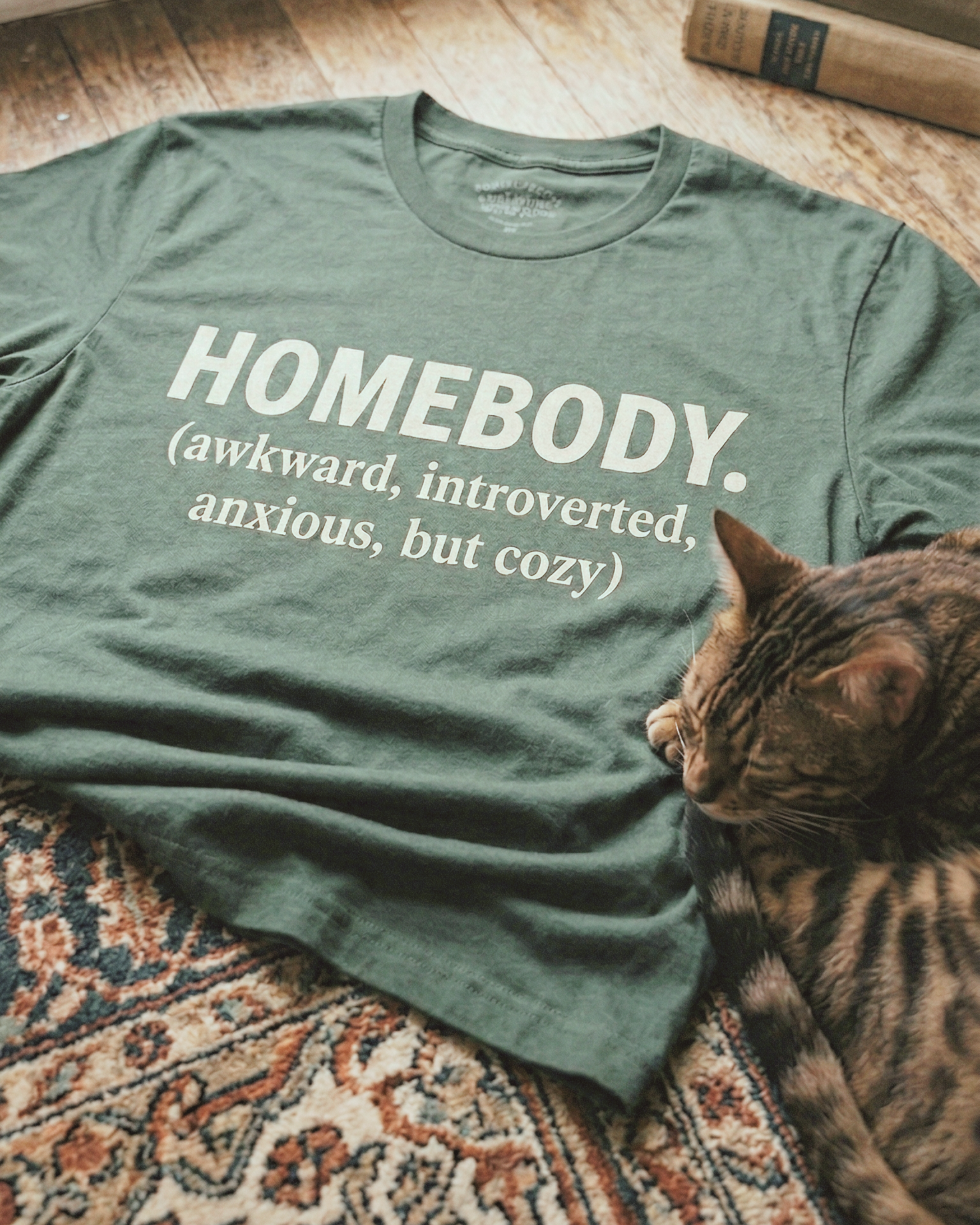 Homebody graphic tee on a wood floor beside a curled up Bengal cat, graphic apparel for introverts, homebodies, and the quietly exhausted
